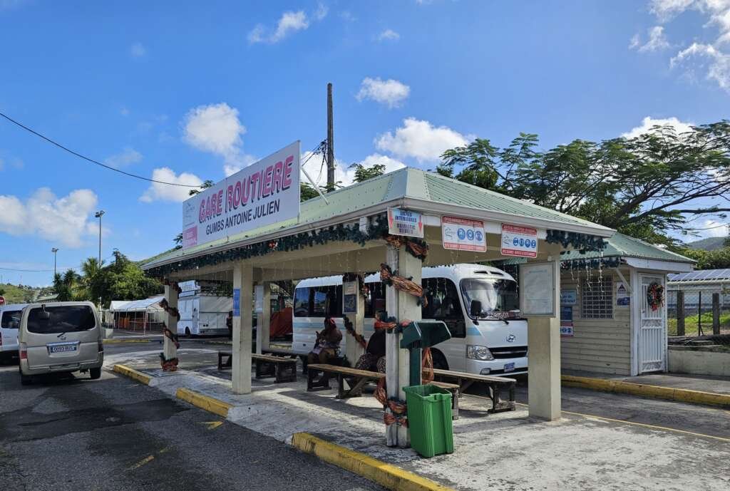 Bus terminal shelter with a large pink GARE ROUTIÈRE sign on the roof and people sitting under the shelter against a blue sky