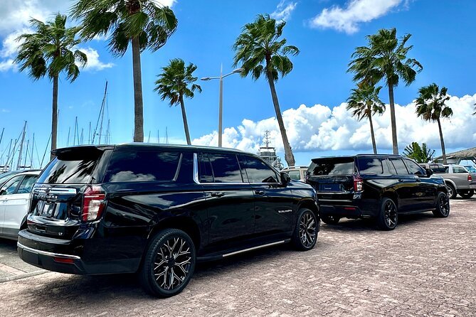 Two black full size SUVs parked on a brick promenade beside a marina with palm trees and a bright blue sky