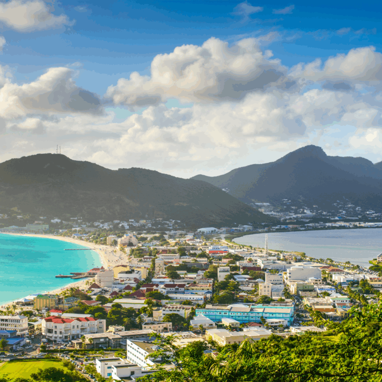 Coastal town with a turquoise bay sandy beach and green hills under a cloudy blue sky at noon