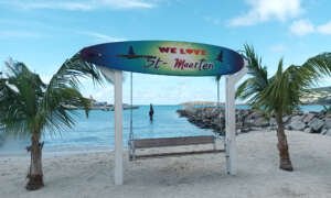 Colorful surfboard sign reading WE LOVE St Maarten above a wooden swing on a sandy beach with palm trees and calm turquoise water behind