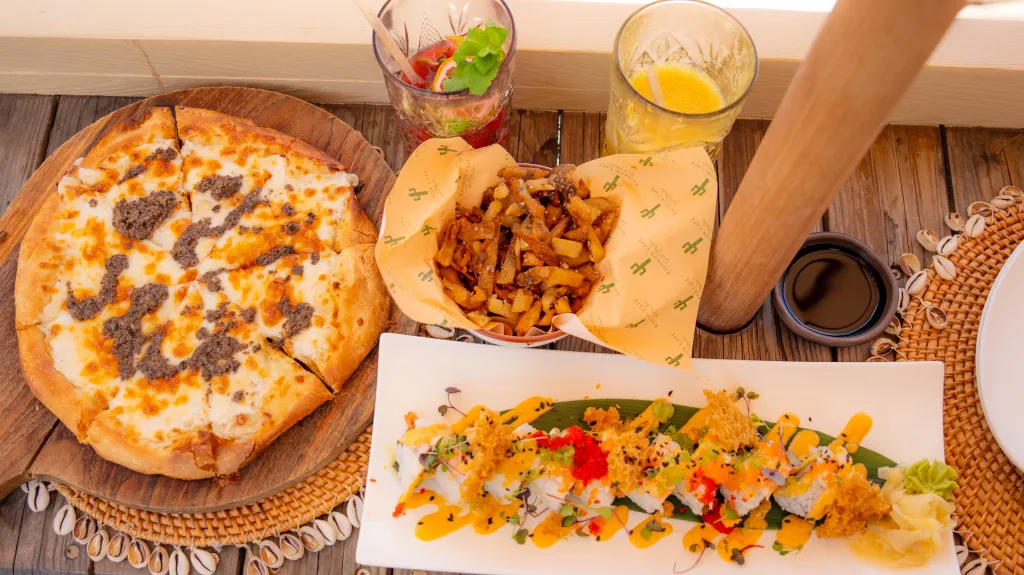 Pizza with melted cheese and toppings on a wooden board beside a bowl of fries and a colorful sushi roll plate row