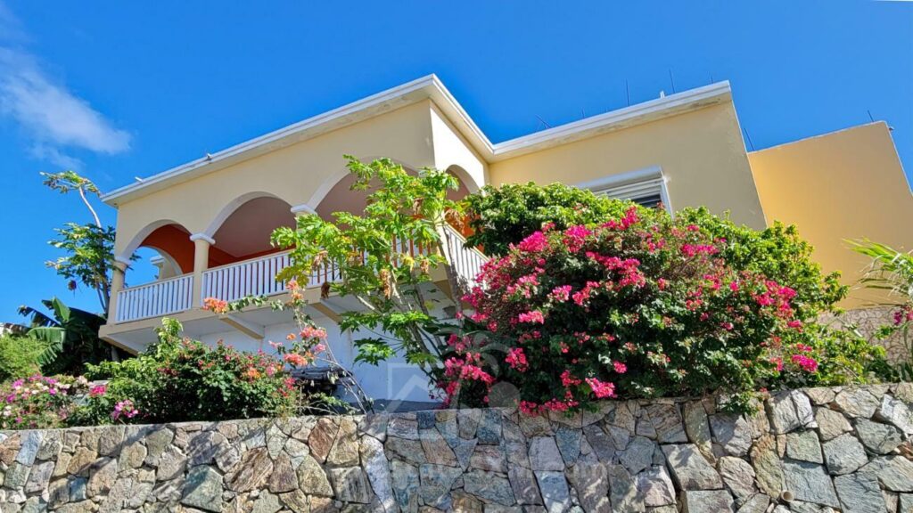 20230908_090345 - Wei Landgraf Two-story Mediterranean-style house with yellow walls, white arches, and a wraparound balcony, set above a stone retaining wall with flowering shrubs.