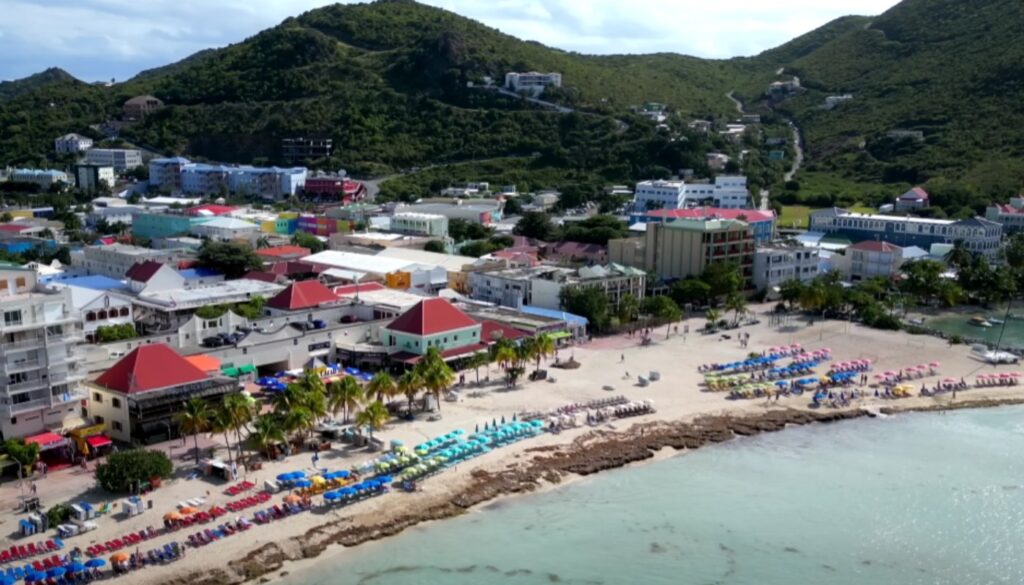 A tropical seaside town with a busy beach lined by colorful umbrellas and palm trees, hills in the background.