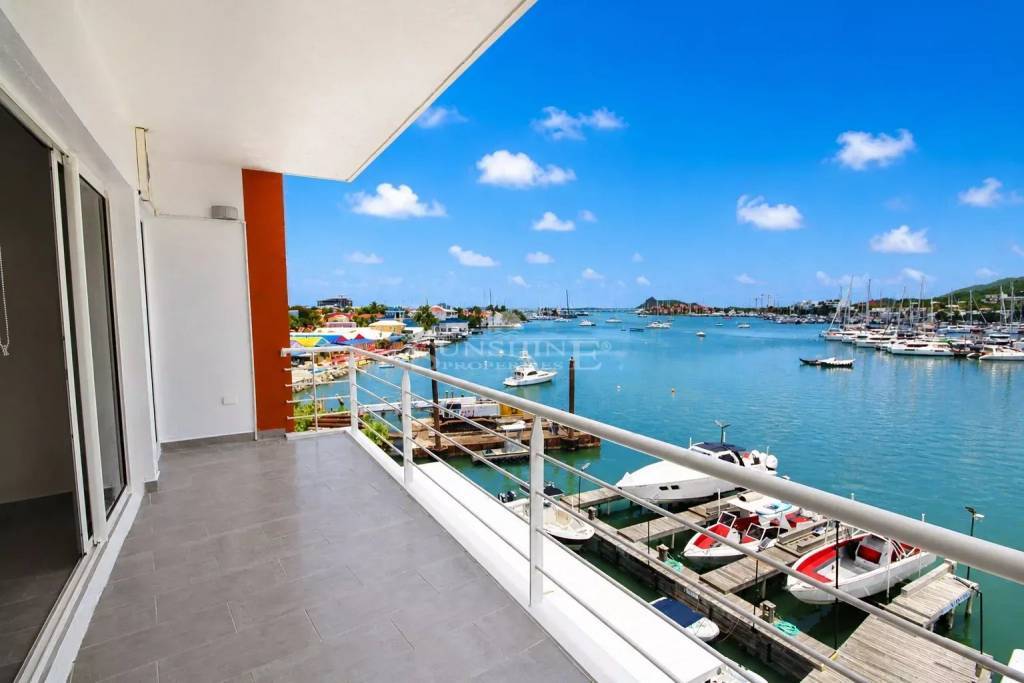 3a91338cff47aa4ec57a45e8ec393172 - Wei Landgraf Balcony overlooking a marina with moored boats, turquoise water, and a bright blue sky; modern railing and tiled floor suggest an apartment or hotel terrace.