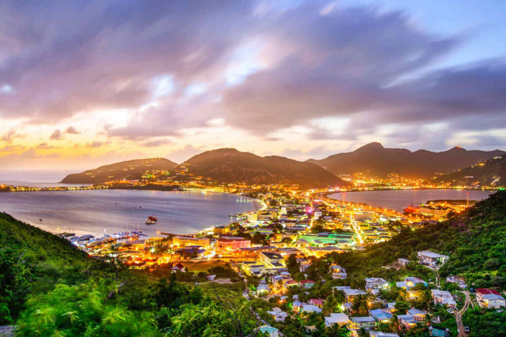 6-Best-places-where-to-stay-in-St-Maarten - Wei Landgraf Evening view of a harbor town along a curved coastline, with illuminated streets and green hills in the foreground and mountains in the background.