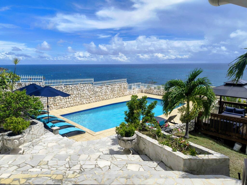 IMG_20250907_133310768_HDR2-scaled-1 - Wei Landgraf Stone terrace with a rectangular blue-tiled pool, blue lounge chairs and umbrellas, overlooking the ocean under a blue sky with clouds, surrounded by palm plants.