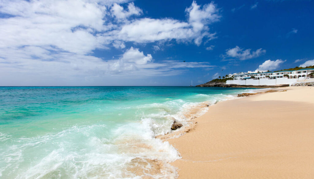 Bright turquoise ocean meets a sandy beach under a clear blue sky, with white villas along the coastline to the right?