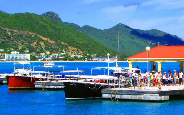 People on a wooden pier beside a red-roofed shelter, with moored boats, calm blue water, and green mountains in the background.