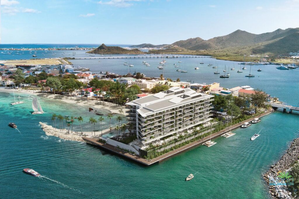 A modern multi-story waterfront hotel on a small peninsula, with palm trees, a sandy beach, and a marina full of boats nearby.