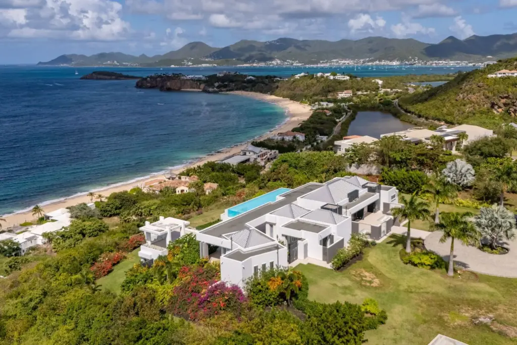 Aerial view of white modern villas along a tropical hillside next to a sandy beach and blue sea.