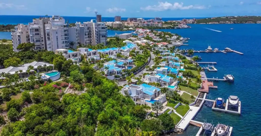 Aerial view of a coastal resort with white buildings, blue-roofed villas, lush greenery, and a marina along bright blue water.