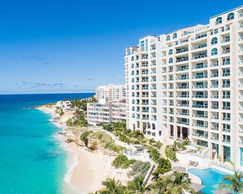 White beachfront hotel complex with balconies along turquoise sea and sandy shore, under a clear blue sky.