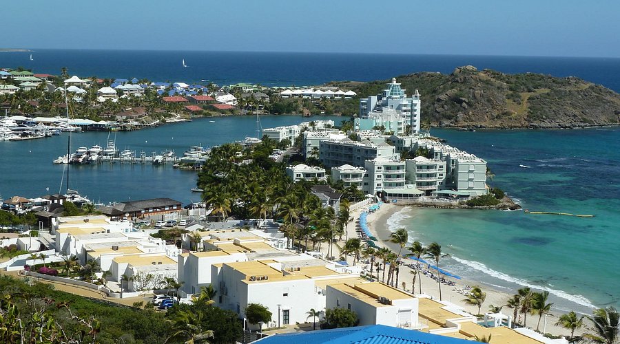part-of-dawn-beach-is - Wei Landgraf Coastal resort town with white buildings, palm trees, a sandy beach, and turquoise water near a marina.