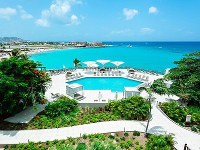 Resort pool area with white lounge chairs, umbrellas, and turquoise sea beyond