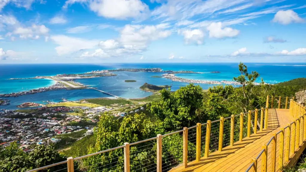 stmaartentourismcovid - Wei Landgraf Wooden railing walkway on a hill overlooking a tropical coastal town and turquoise bay under a bright blue sky.