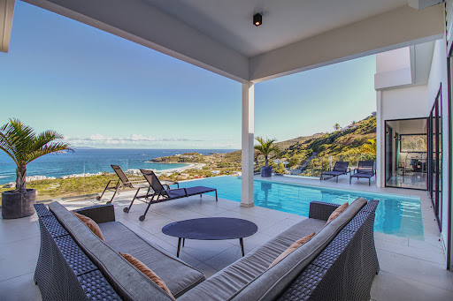unnamed-2026-05-26T164414309 - Wei Landgraf Covered outdoor patio with wicker seating around a round table, a pool, and lounge chairs overlooking the ocean and rocky coastline.