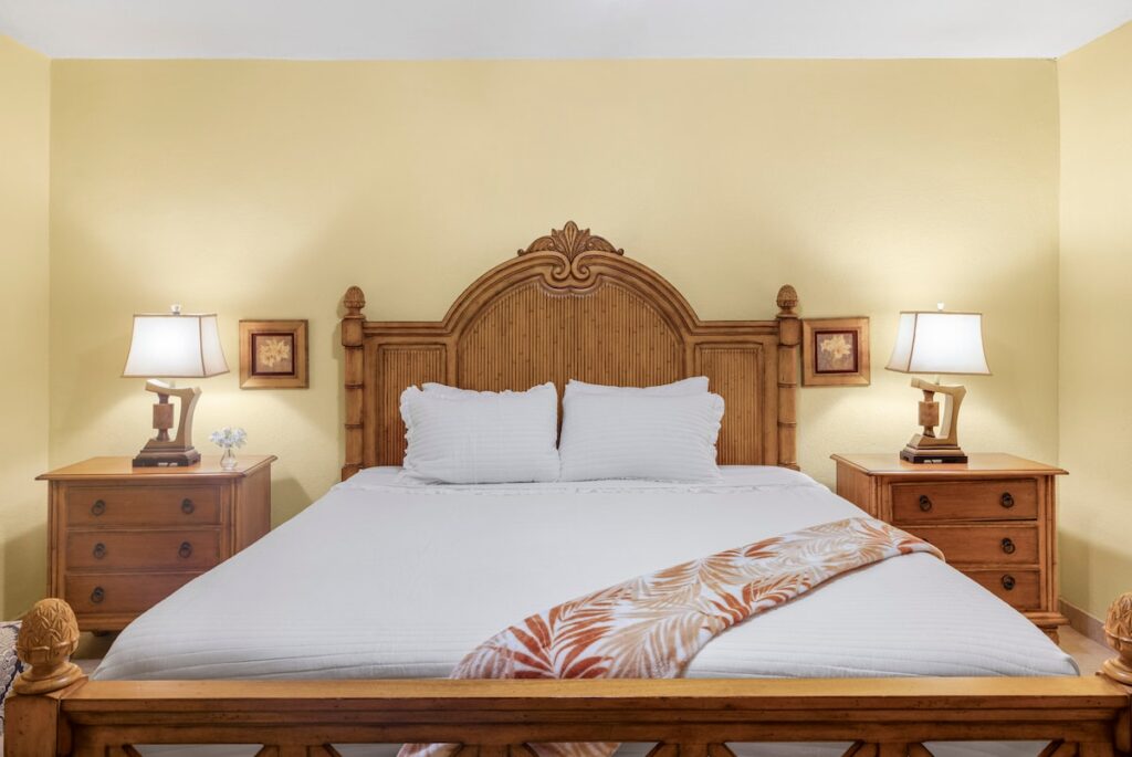 Wooden bed with an ornate carved headboard, white bedding, and an orange palm-pattern throw, flanked by two nightstands with lamps in a yellow-toned bedroom.