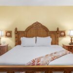 Wooden bed with an ornate carved headboard, white bedding, and an orange palm-pattern throw, flanked by two nightstands with lamps in a yellow-toned bedroom.