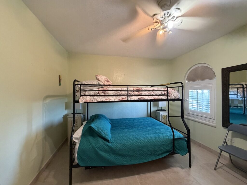 Small bedroom with a black metal bunk bed: teal quilt on bottom bed and pink floral quilt on top, pale green walls, ceiling fan, window with blinds, and a folding chair nearby.