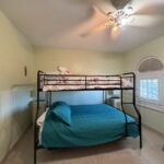 Small bedroom with a black metal bunk bed: teal quilt on bottom bed and pink floral quilt on top, pale green walls, ceiling fan, window with blinds, and a folding chair nearby.