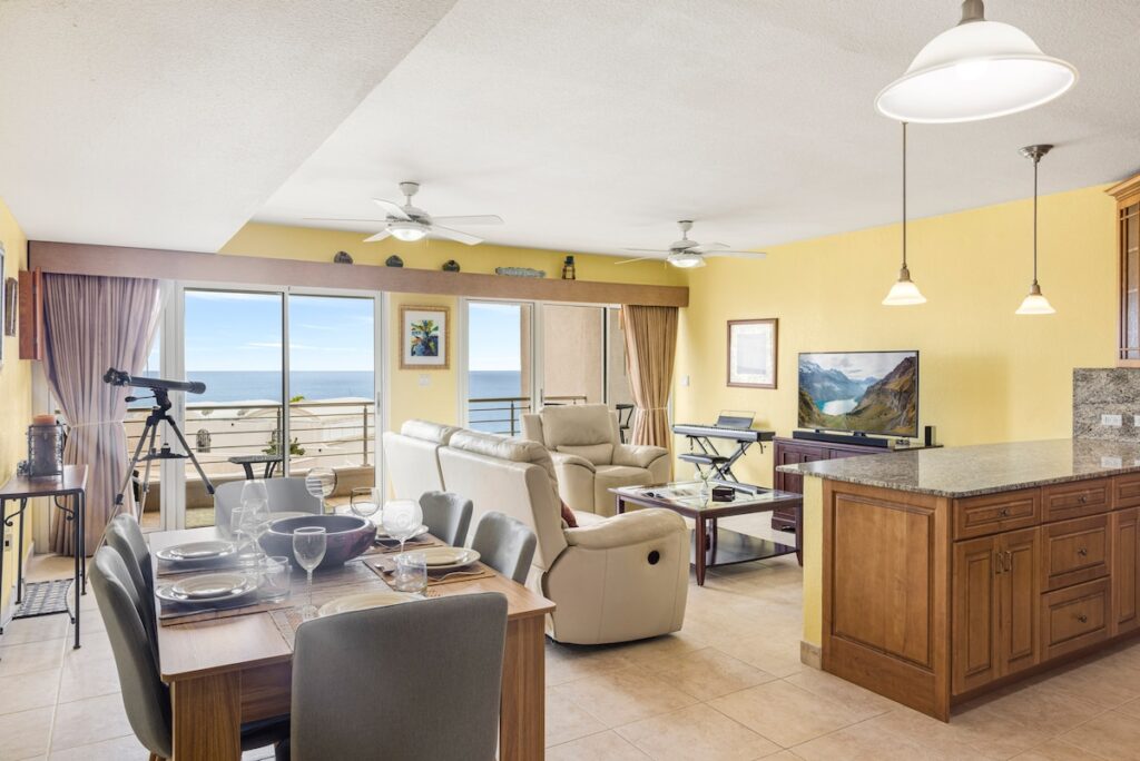 Open living and dining area with beige reclining sofa, dining table for six, and a balcony view of the sea beyond sliding glass doors to the right.
