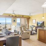 Open living and dining area with beige reclining sofa, dining table for six, and a balcony view of the sea beyond sliding glass doors to the right.