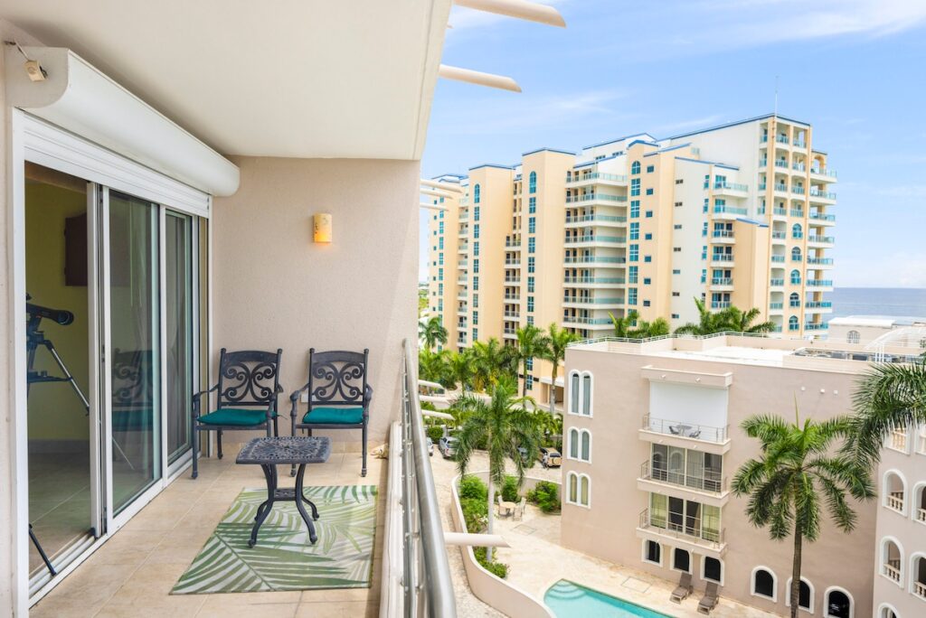 Balcony with two ornate chairs and a small table, overlooking a resort-style complex with tall beige buildings and palm trees, ocean in distance