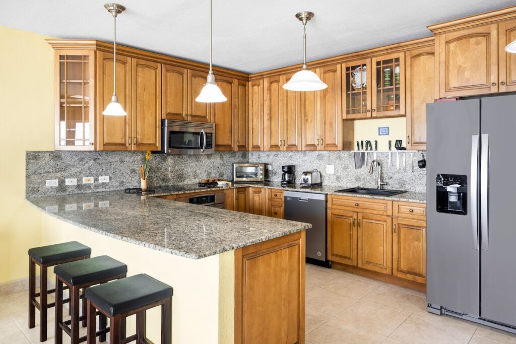 Bright kitchen with granite countertops, wooden cabinets, and an island with three stools beneath pendant lights.