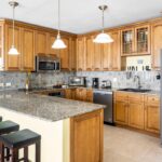 Bright kitchen with granite countertops, wooden cabinets, and an island with three stools beneath pendant lights.