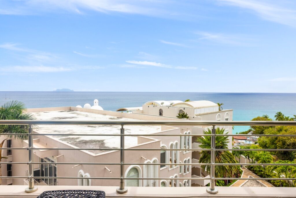 Balcony railing frames beige Mediterranean buildings overlooking a blue sea with a distant island on the horizon.