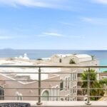 Balcony railing frames beige Mediterranean buildings overlooking a blue sea with a distant island on the horizon.