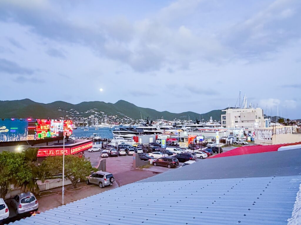 Marina at dusk with parked cars along a promenade, colorful neon signs, and moored boats with hills in the background...