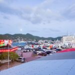 Marina at dusk with parked cars along a promenade, colorful neon signs, and moored boats with hills in the background...