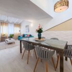 Bright open-plan living and dining area with a glass-top table, gray chairs, and a blue sofa under a sloped ceiling.",