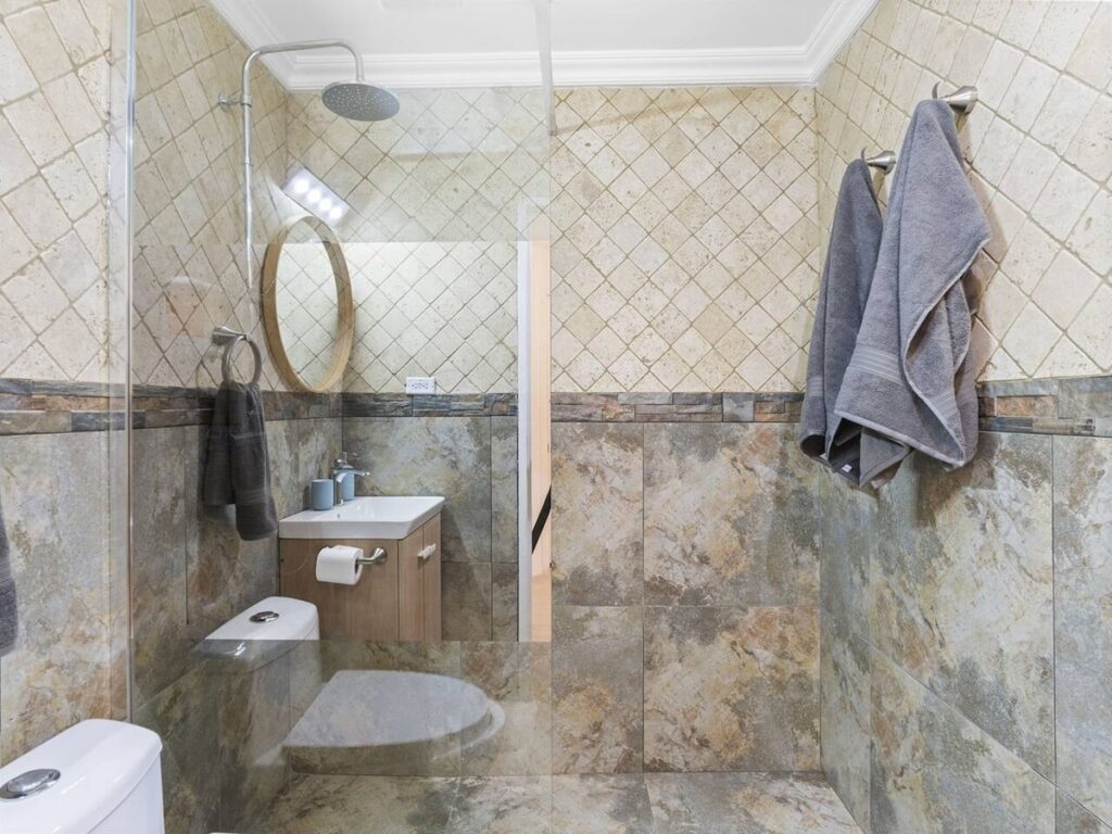 Modern bathroom with glass shower enclosure, beige marbled tiles, and gray towels hanging on the wall rack.