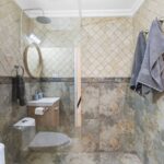 Modern bathroom with glass shower enclosure, beige marbled tiles, and gray towels hanging on the wall rack.