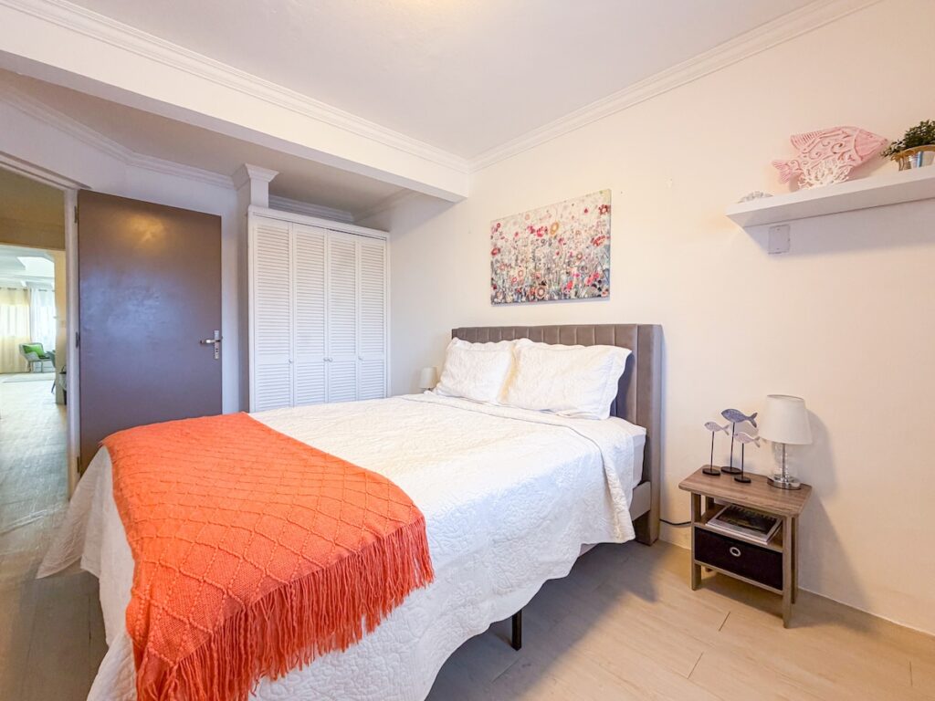 Bright bedroom with white quilted bed, orange throw, and gray tufted headboard; bedside table with lamp and decor on the right.