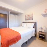 Bright bedroom with white quilted bed, orange throw, and gray tufted headboard; bedside table with lamp and decor on the right.