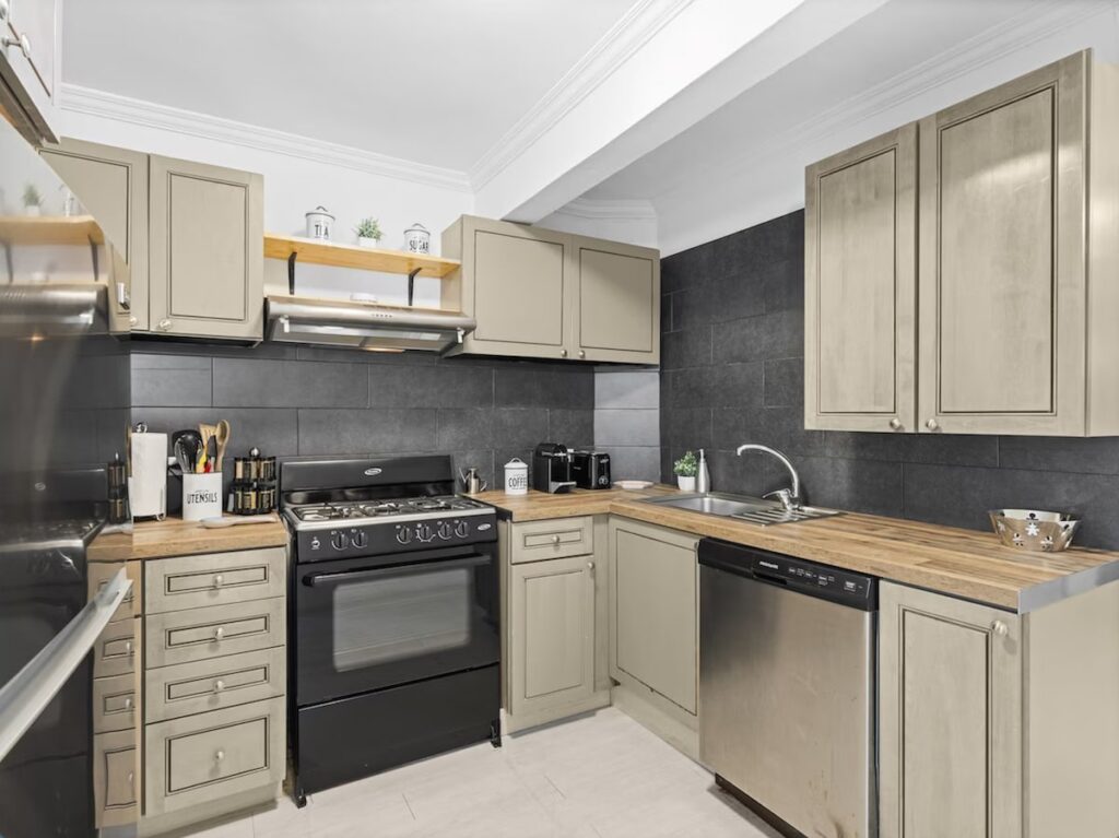 L-shaped kitchen with beige raised-panel cabinets, black gas range, stainless dishwasher, and wooden countertops against a dark tile backsplash.