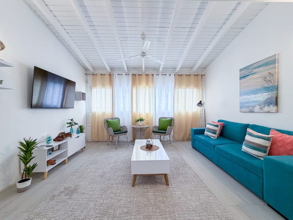 Bright coastal living room with teal sofa, two wicker chairs, white coffee table, beige curtains, and a beach scene painting.