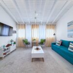 Bright coastal living room with teal sofa, two wicker chairs, white coffee table, beige curtains, and a beach scene painting.