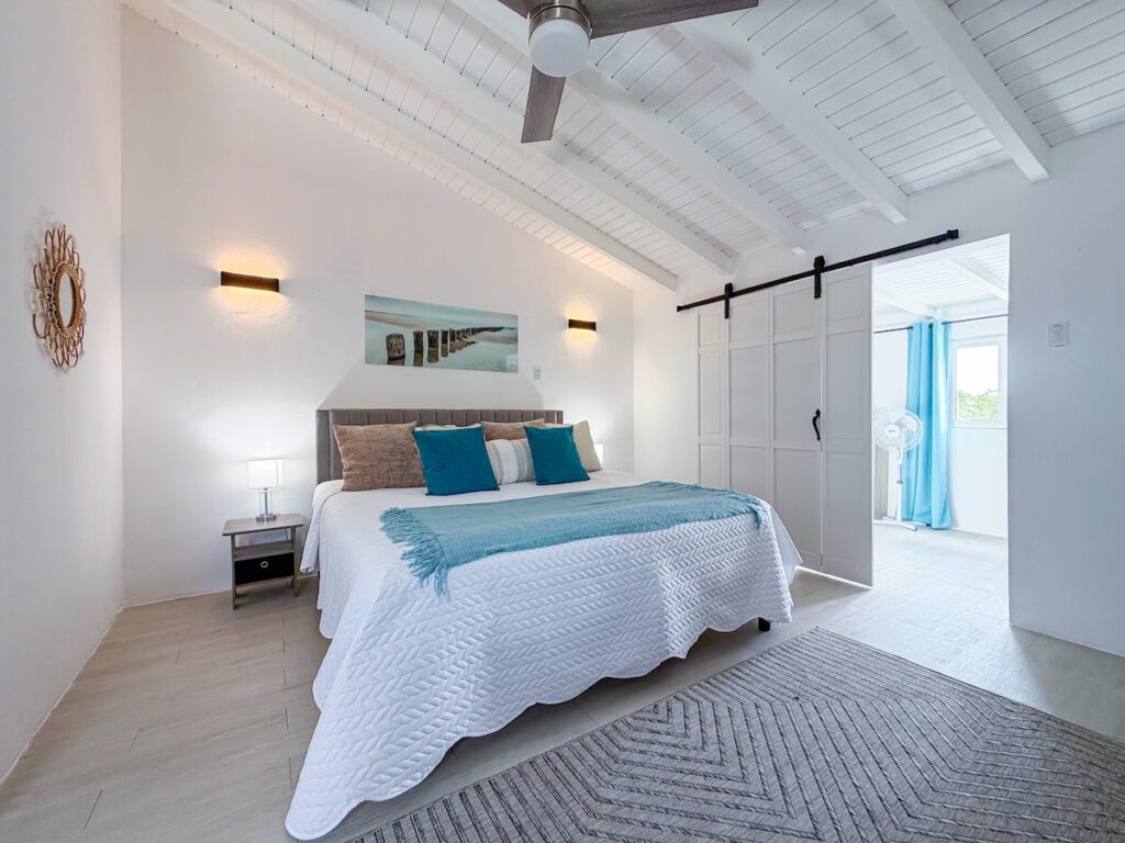 Bright white bedroom with vaulted, beamed ceiling; bed with white quilt, blue pillows, and a light blue throw.