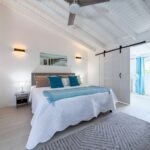 Bright white bedroom with vaulted, beamed ceiling; bed with white quilt, blue pillows, and a light blue throw.